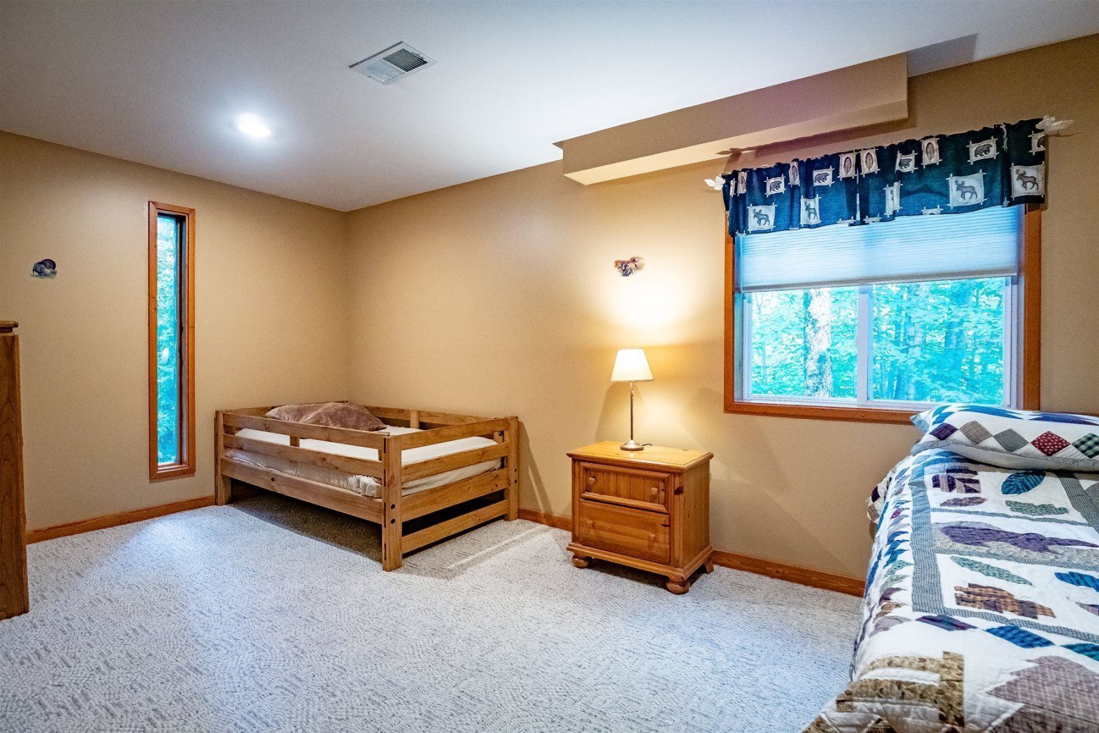 A bedroom with a bunk bed, a bedside table with a lamp, a window with a view of trees, and a quilted bedspread.