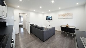 A modern living room with a grey sofa, a dining table with two chairs, a flat-screen TV mounted on the wall, and a kitchen area with a stove. - The Hatton Apartments (Feltham)