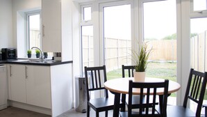 A modern kitchen with white cabinets, a black countertop, and a sink. There is a dining area with a round table and four chairs. A potted plant is on the table. - The Hatton Apartments (Feltham)