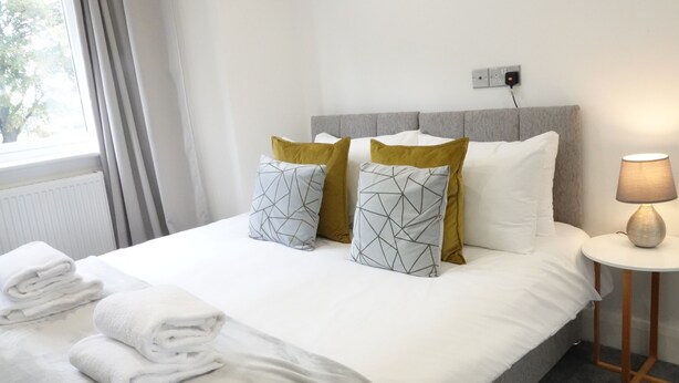 A neatly made bed with white bedding and geometric patterned pillows, a bedside lamp, and a window with curtains. - The Hatton Apartments (Feltham)