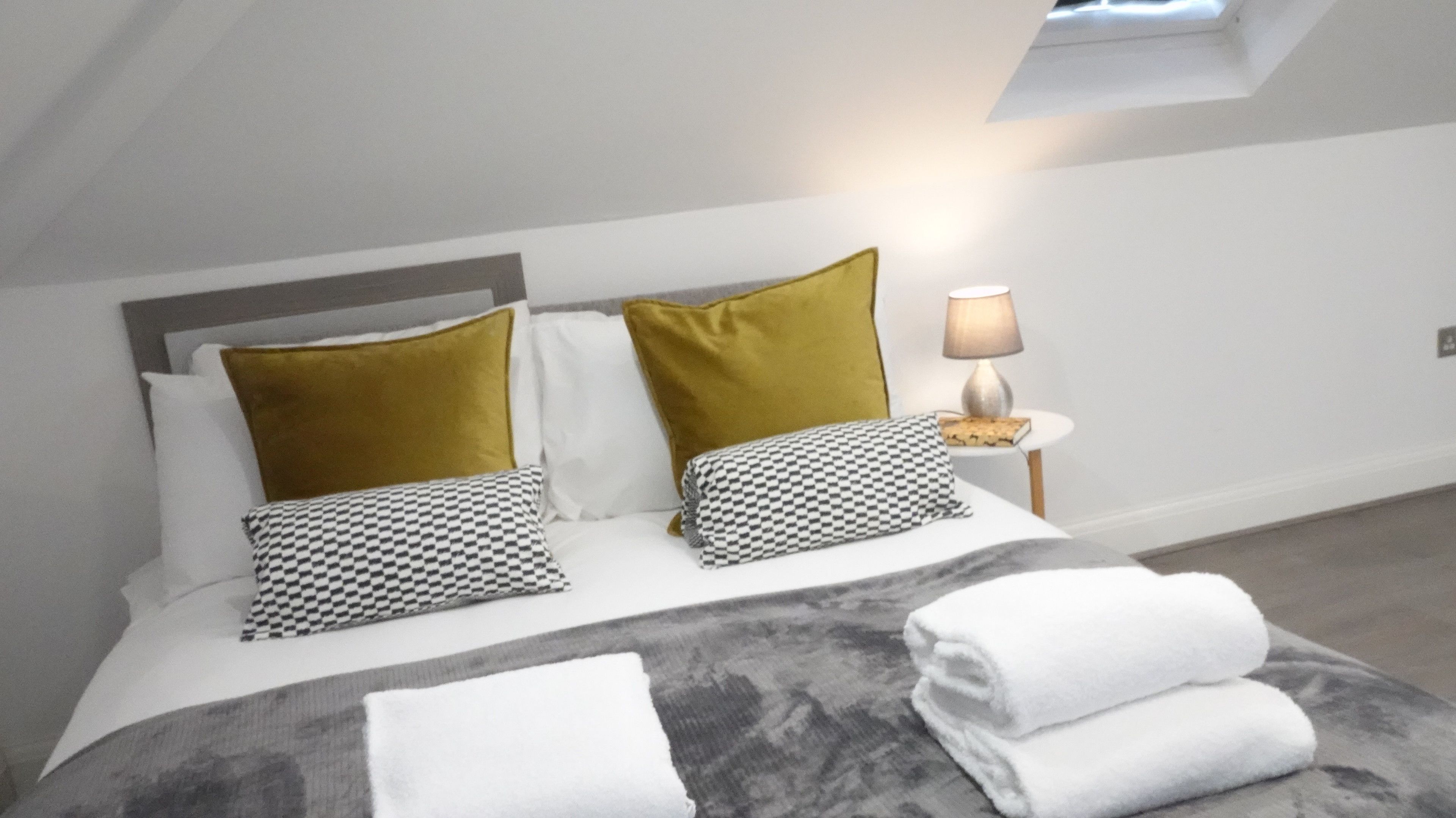 Una cama bien hecha con un almohadón gris y blanco a cuadros, un almohadón blanco y un almohadón color mostaza. Hay una lámpara sobre una mesita de noche junto a la cama.