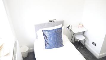 A neatly arranged hotel room with a bed, a small desk, a chair, a lamp, and a trash bin.