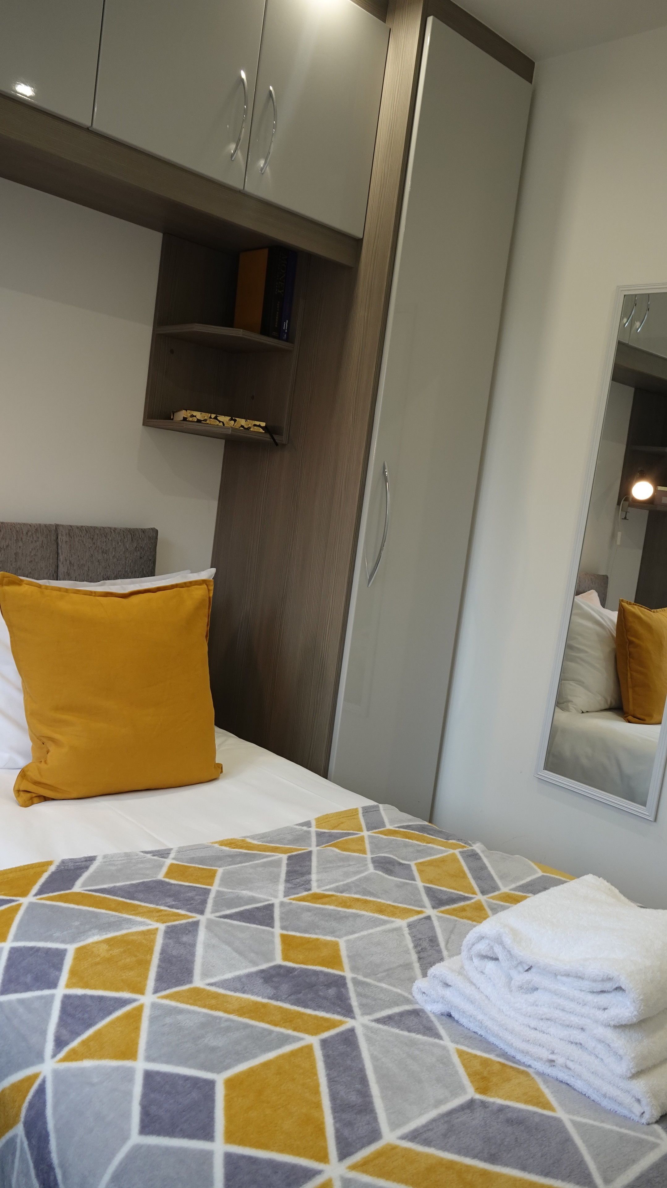 Quarto de hotel com uma cama, guarda-roupa, espelho e uma estante com livros.