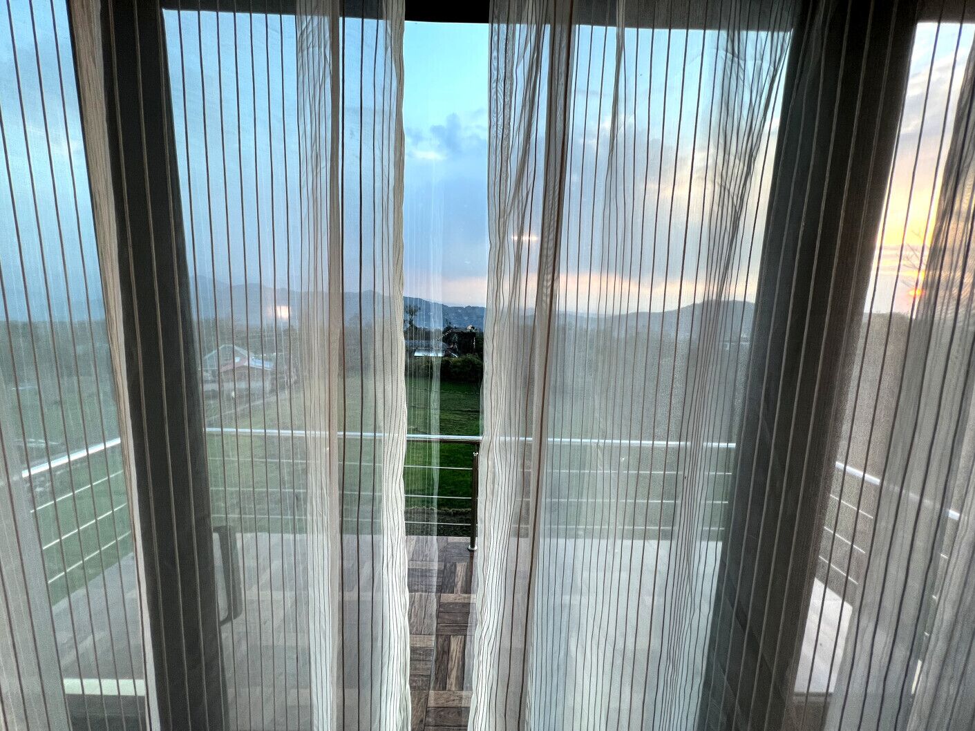 Vista a través de cortinas translúcidas, mostrando un estacionamiento y un paisaje lejano con montañas.