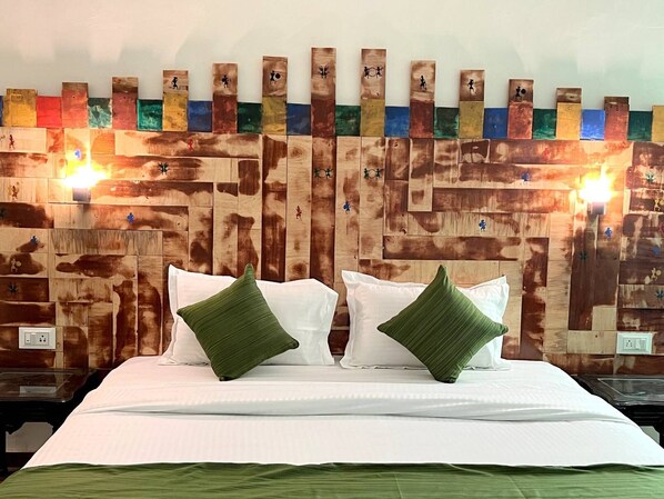 Una cama con ropa blanca y dos almohadas verdes, cabecera de madera con un mural colorido, y dos lámparas fijadas a la pared.