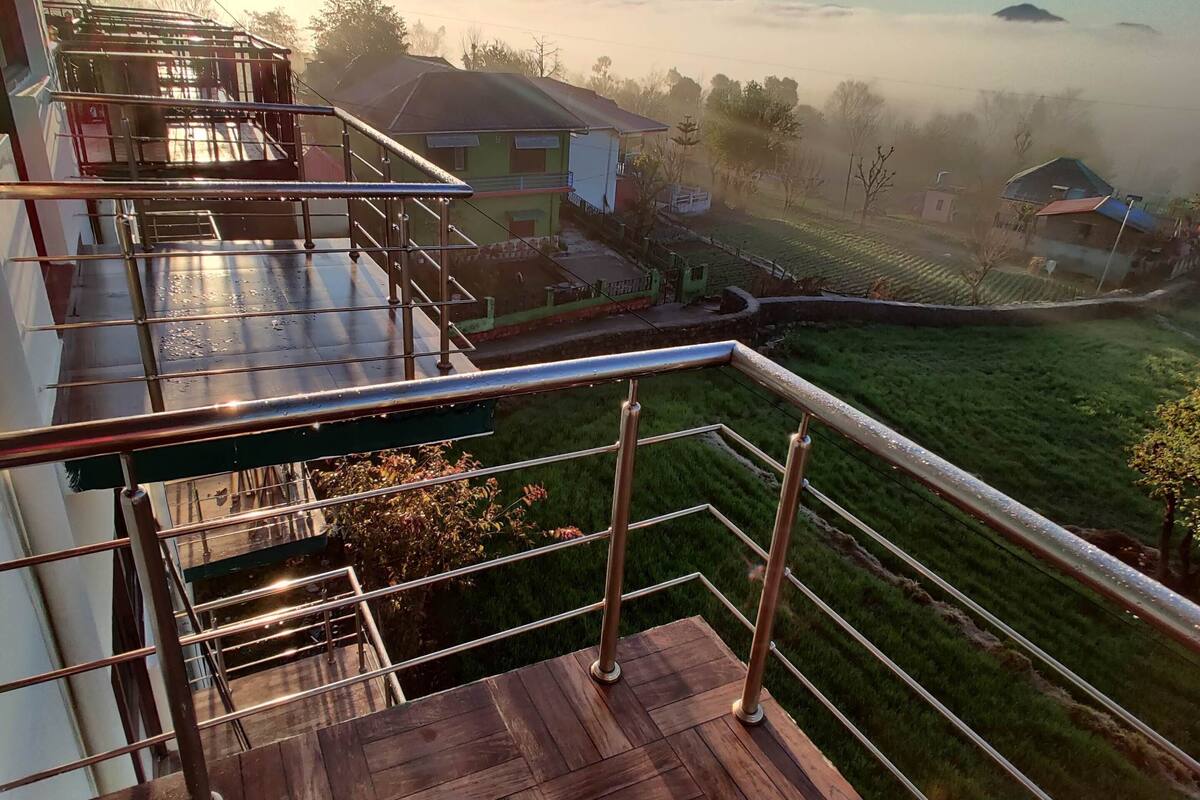 Een houten terras met metalen hekken met uitzicht op een landelijk landschap met huizen en een mistige berg in de verte.