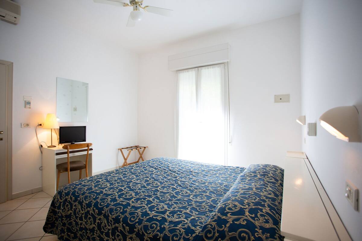 A bedroom with a bed, a desk with a computer, a chair, a ceiling fan, and a window with curtains.