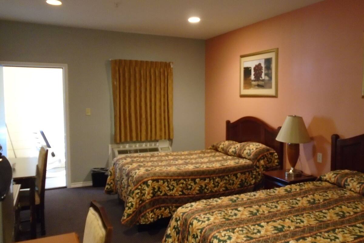 Hotelkamer met twee bedden, een bureau, een stoel, een raam met gordijnen, een airconditioner, een lamp en een lijst aan de muur.