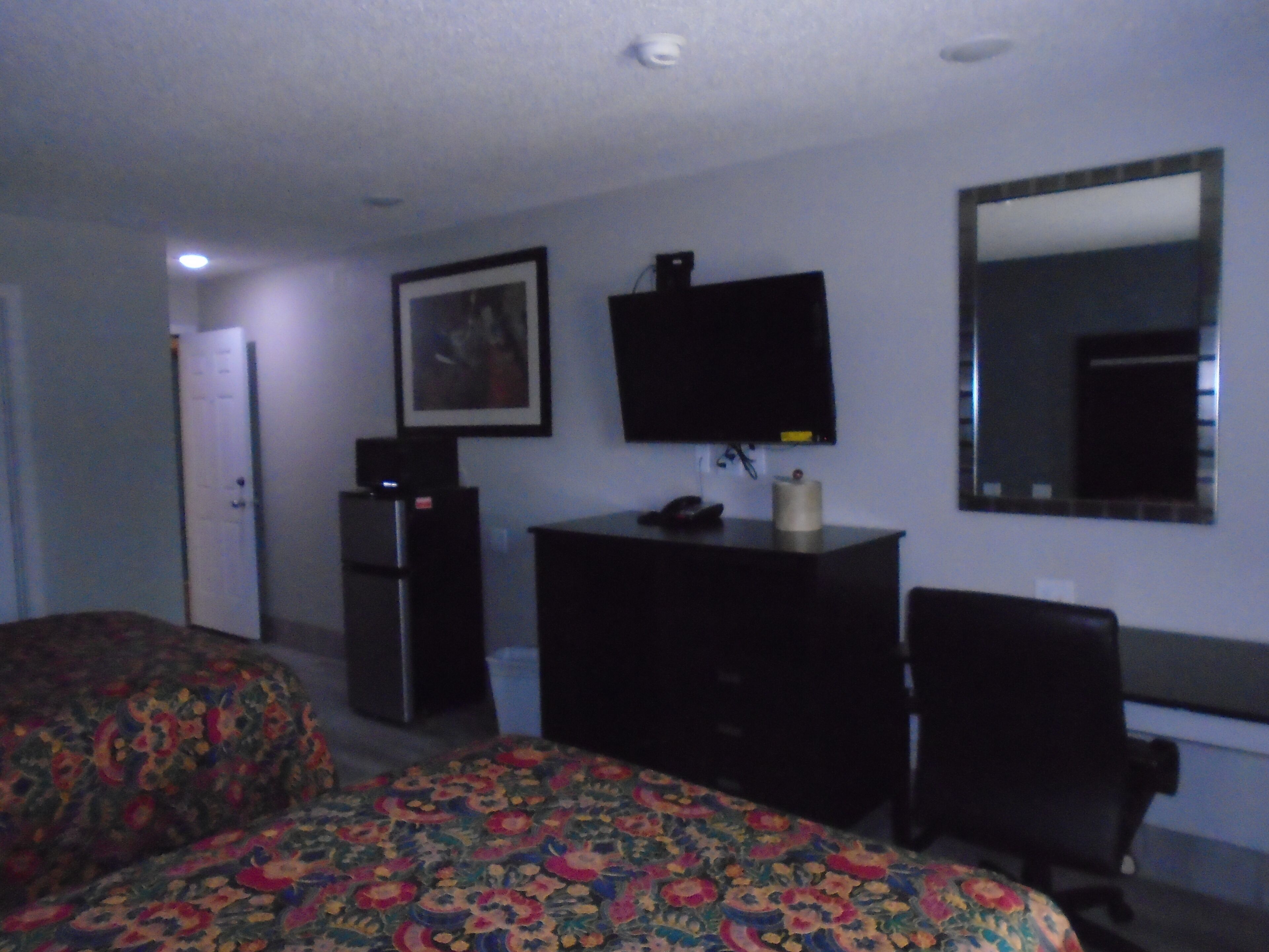 Quarto de hotel com cama, escrivaninha, cadeira, televisão e espelho.