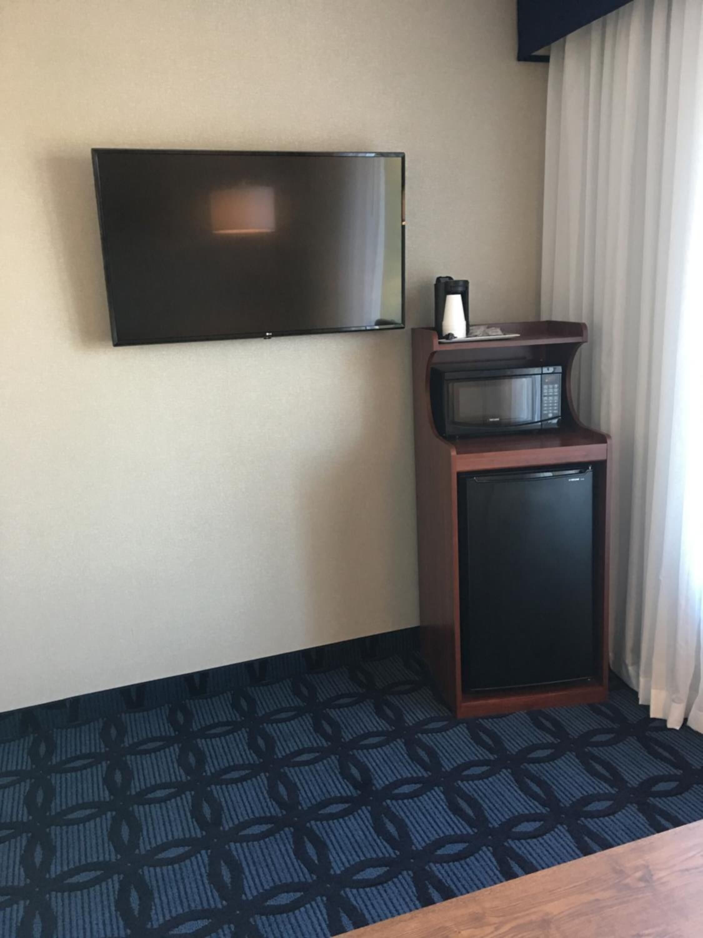 A hotel room with a flat-screen TV mounted on the wall, a microwave on a small wooden cabinet, and a patterned carpet.
