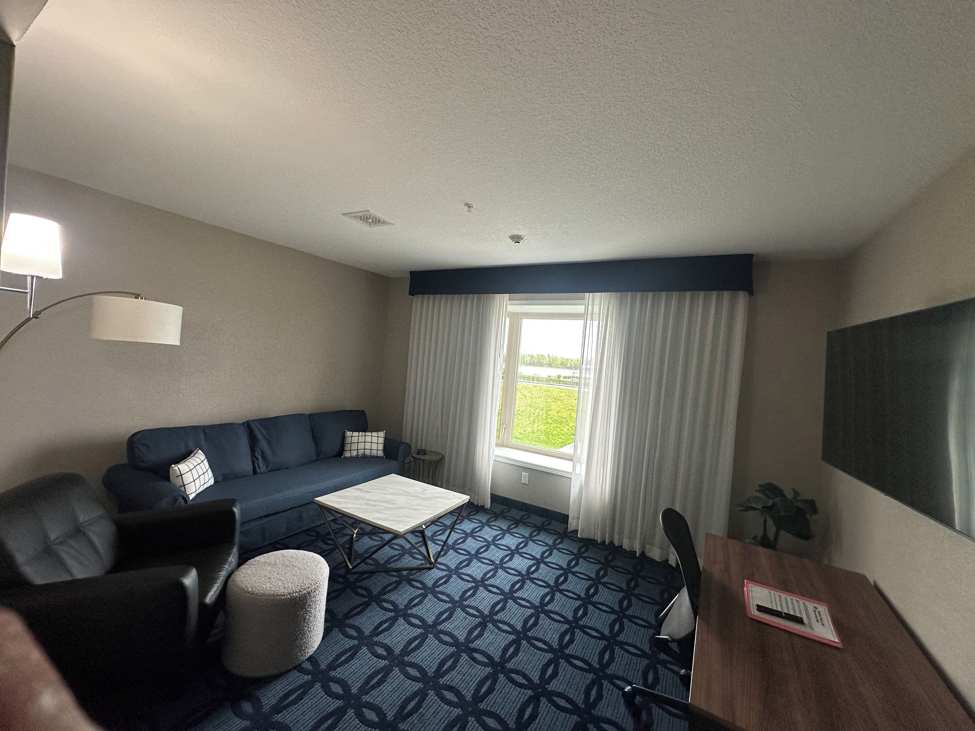 Hotelkamer met blauw behang met patroon, een blauwe bank, een klein rond voetenbankje, een houten bijzettafeltje, een lamp, een raam met gordijnen en uitzicht op groen.