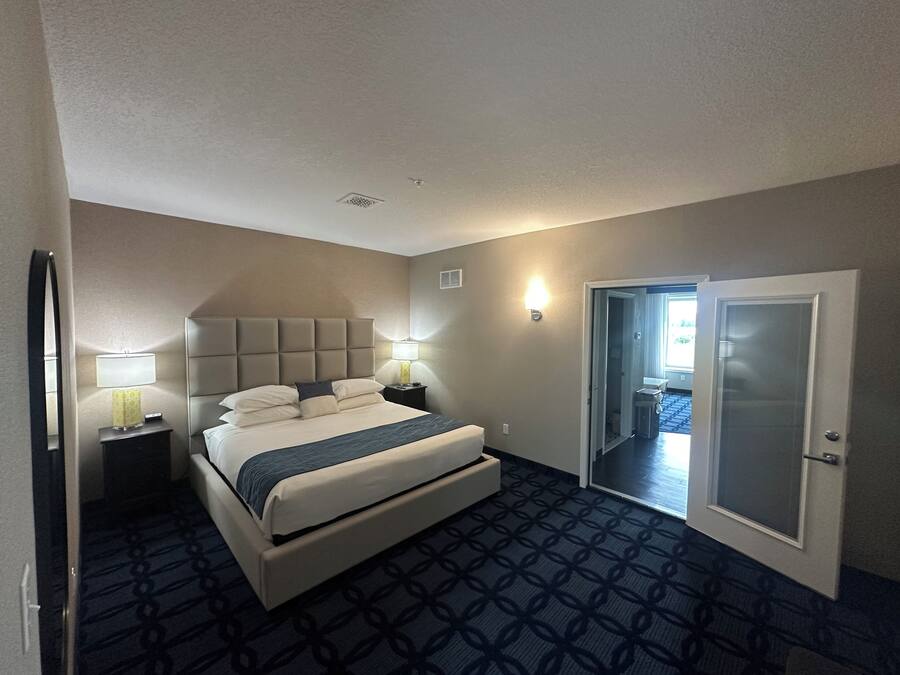 Habitación de hotel con una cama, dos mesitas de noche, una lámpara, un espejo y una puerta que da a otra habitación.