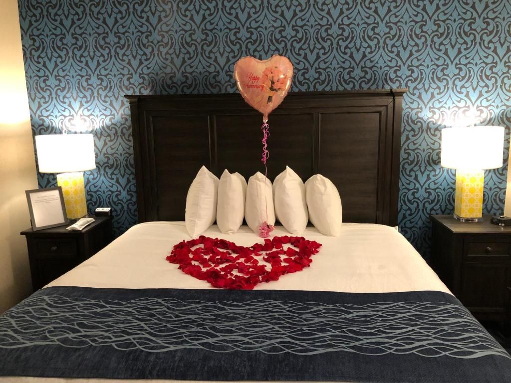 A hotel room with a bed, a heart-shaped arrangement of rose petals, a heart-shaped balloon, and a patterned wall.