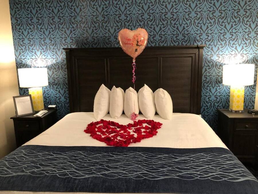A hotel room with a bed, a heart-shaped arrangement of rose petals, a heart-shaped balloon, and a patterned wall.