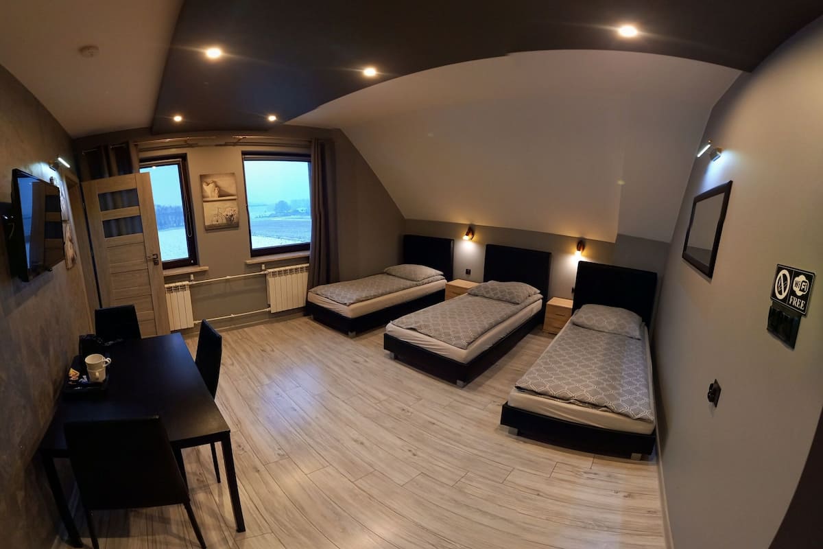 A compact hotel room with three single beds, a dining table, and a view of the sea through a large window.