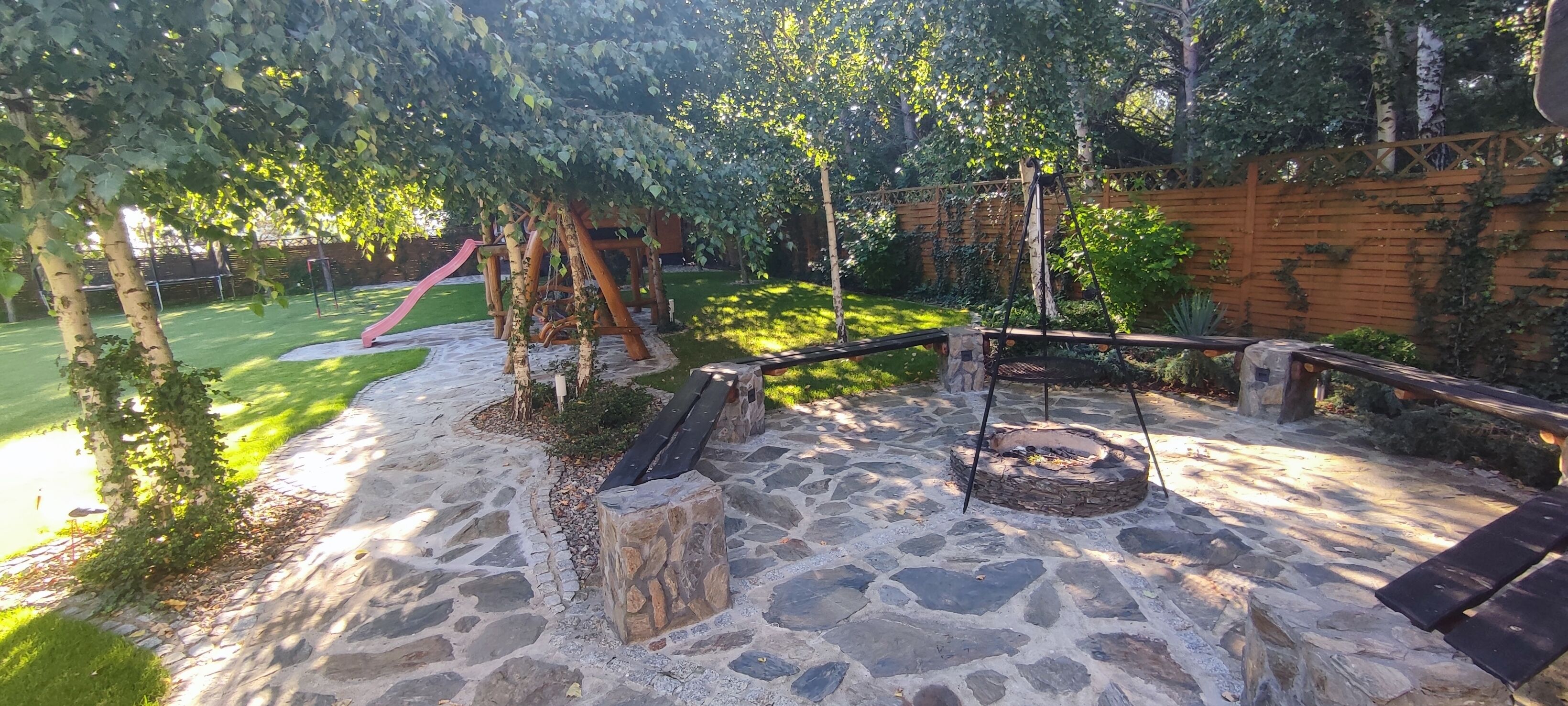 Un patio de piedra con un brasero, bancos de madera y un conjunto de columpios en un jardín.