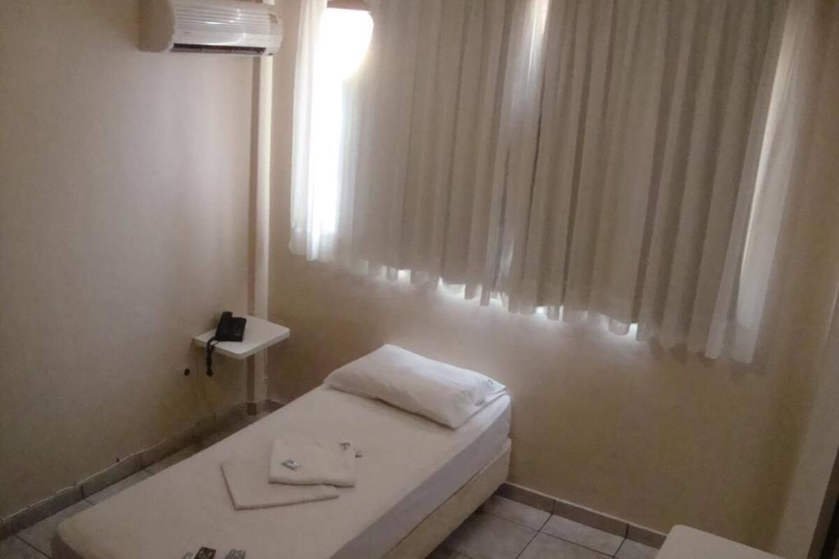 A single bed with a white mattress and pillow, a bedside table with a phone, and a window with curtains.