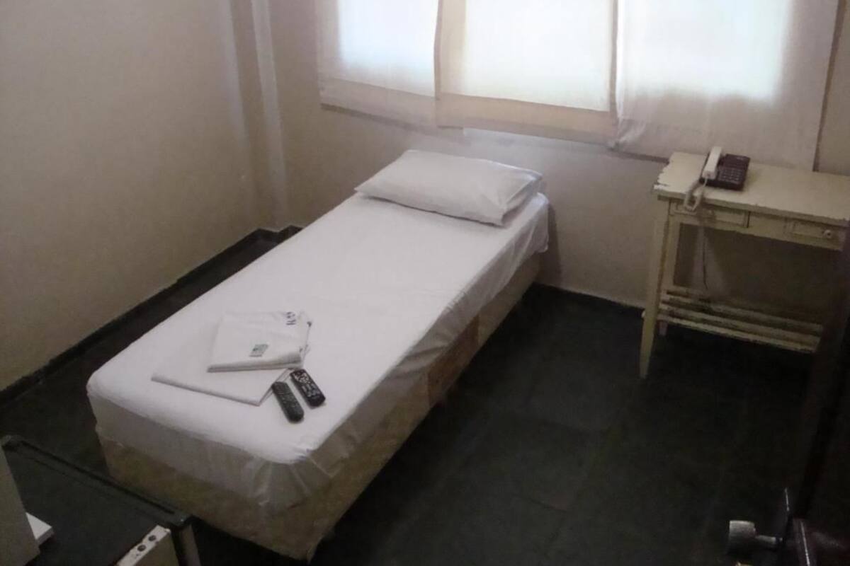 A small, single-bed room with a white mattress and pillow, a bedside table with a telephone, and a window with sheer curtains.