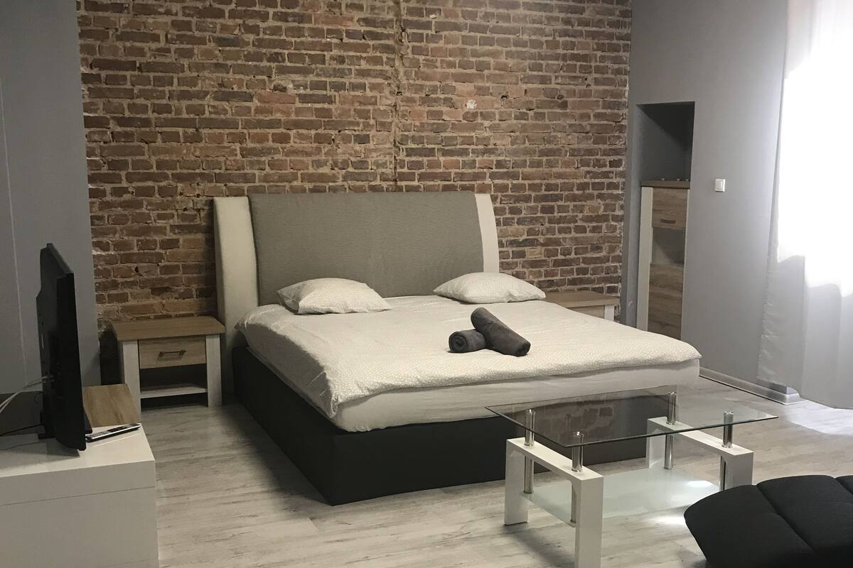 A modern bedroom with a brick wall, a large bed, a glass coffee table, and a black sofa.