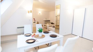 A modern dining area with a white table set for four, a small plant centerpiece, and a view into a living space with a sofa and a coffee table. - Studio Apartament Centrum Katowice (Katowice)