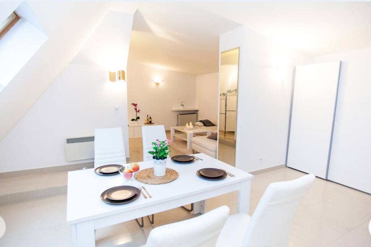 A modern dining area with a white table set for four, a small plant centerpiece, and a view into a living space with a sofa and a coffee table.