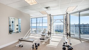 Une salle de sport équipée de matériel d’exercice, d’un banc, d  ’haltères et de grandes fenêtres offrant une vue sur des immeubles et une plage.