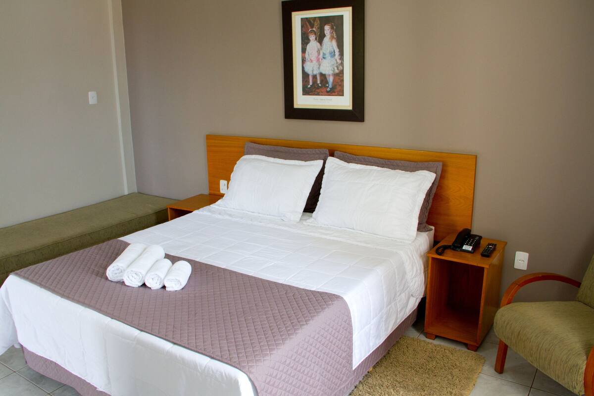 Una cama bien hecha con cubrecolcha acolchado, un cuadro en la pared, una mesita de noche con teléfono y control remoto, y una silla en la esquina.