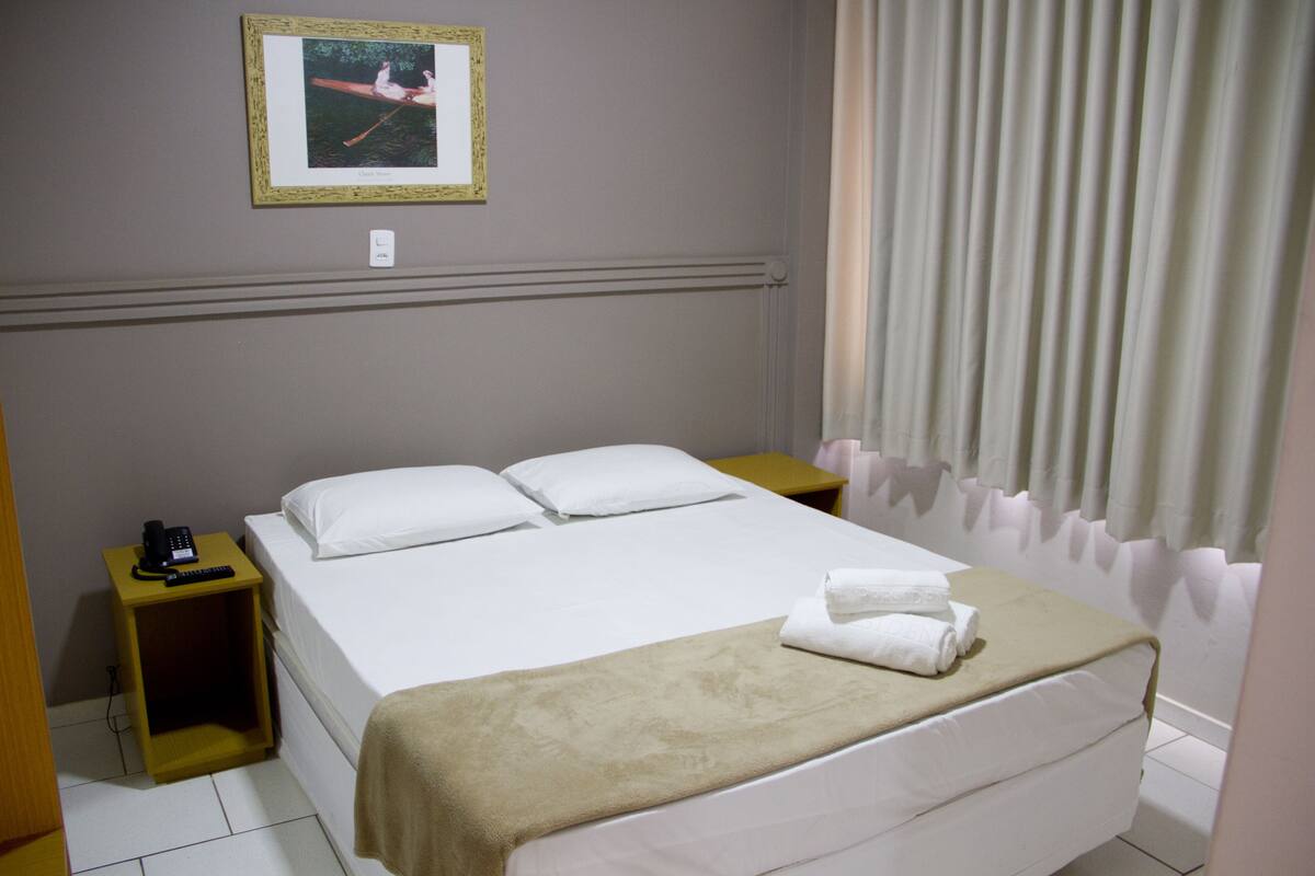 Habitación de hotel con cama, mesita de noche con teléfono, un cuadro en la pared y una ventana con cortinas.