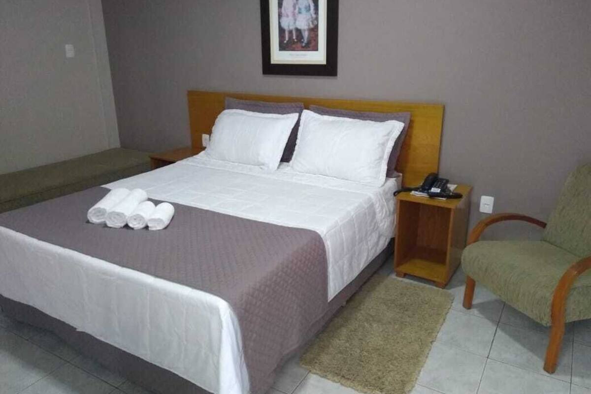 Una cama bien tendida con sábanas blancas, cabecera de madera, mesita de noche con un teléfono y una silla verde.