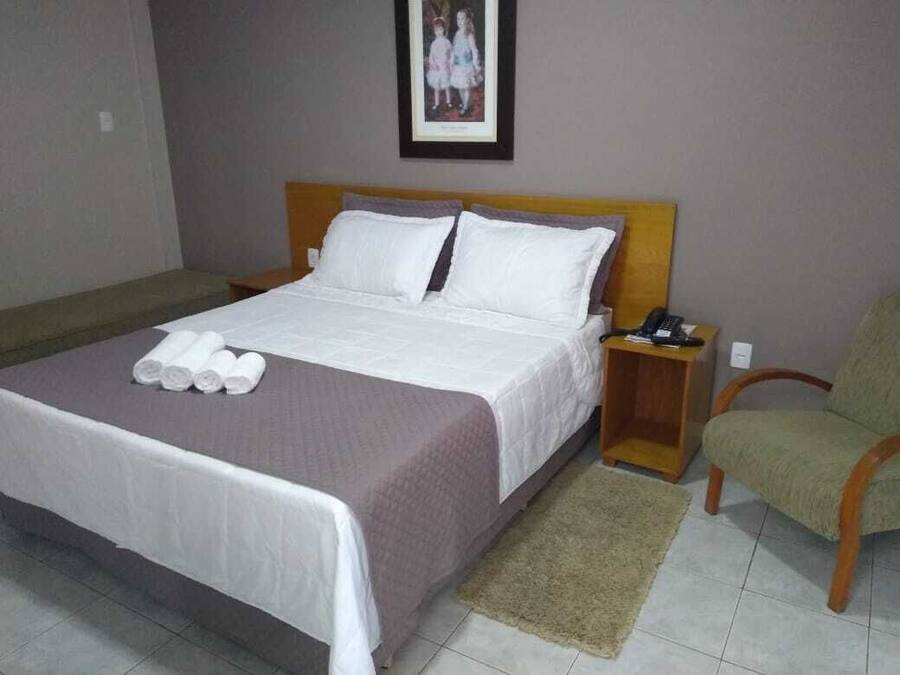 Una cama bien hecha con sábanas blancas, un cabecero de madera, una mesita de noche con un teléfono y una silla verde.