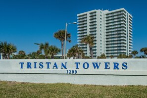Placa com a inscrição 'TRISTAN TOWERS' e o número 1200, rodeada por palmeiras e um edifício alto ao fundo.