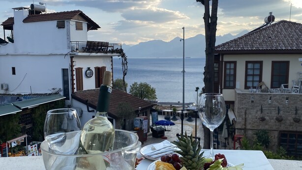 A table with a cheese platter and a glass of white wine, overlooking a coastal view with buildings and mountains. - Mono Hotel (Antalya)