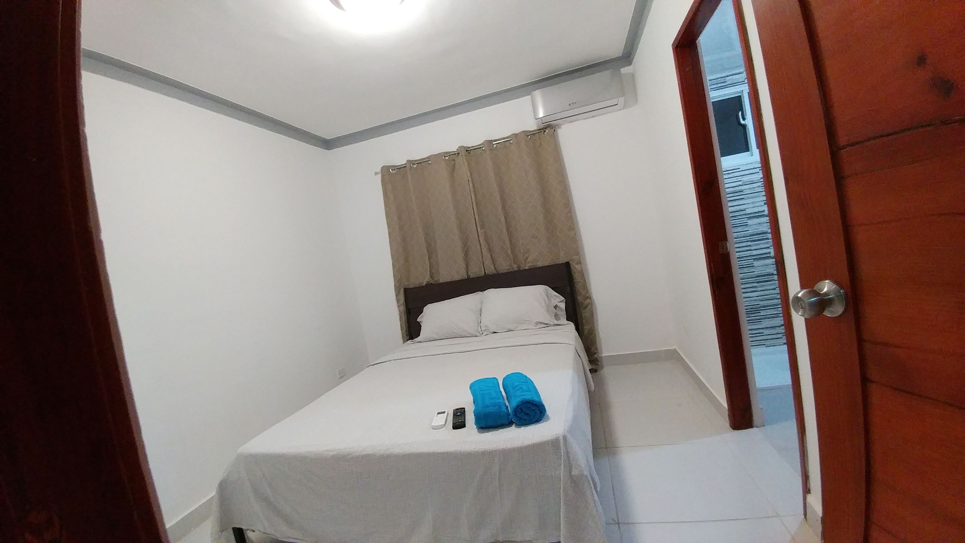 A small, single-bed room with a white bedspread, a remote control, and a rolled-up blue towel on the bed. - Tropical Condo BBQ Pool 5min to Airport (Puñal)