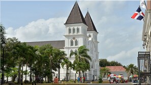 A white church with a tall tower and pointed roof, surrounded by palm trees and a clear sky.