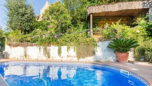 Una piscina con borde de azulejos, rodeada de abundante vegetación y una estructura de techo de paja.