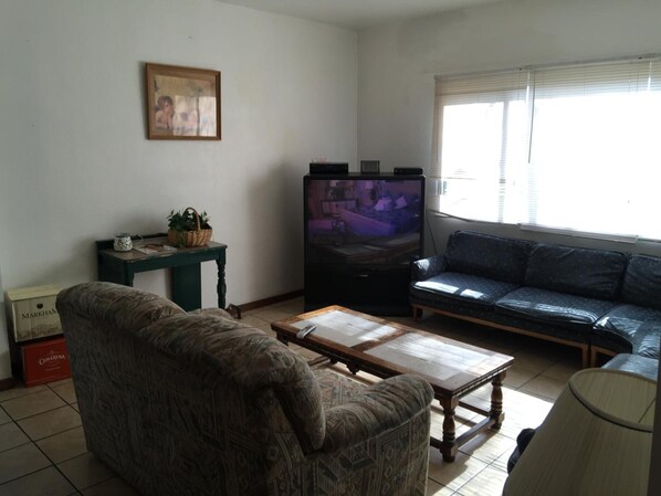 A living room with a patterned armchair, a wooden coffee table, a television, a sofa, and a window with blinds. - Avenues Hostel (Salt Lake City)