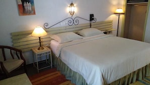 A neatly made bed with a headboard, two bedside lamps, a small table with a lamp, a chair, and a painting on the wall. - Hotel Royal (Oujda)