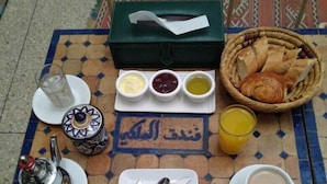 A breakfast setup with a teapot, cups, a basket of bread, and various condiments on a tiled table. - Hotel Royal (Oujda)