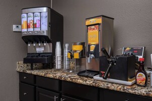 Una estación de café con máquina de café, dispensador de siropes, plancha para gofres y una botella de sirope sin azúcar.