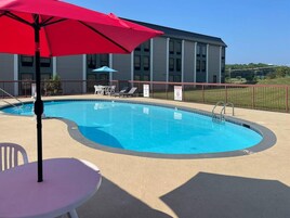 Una piscina con sillas de descanso y sombrillas, una mesa y un edificio al fondo.