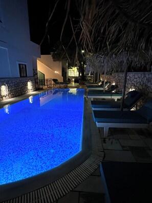 A swimming pool with a blue light, lounge chairs, and a stone wall. - Santa Magia (Santorini)