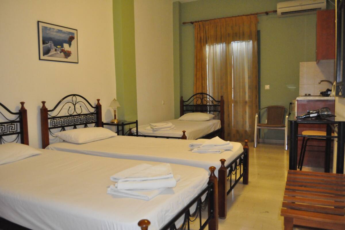 Une chambre d’hôtel avec trois lits simples, un bureau avec une chaise et une salle de bain visible à travers un rideau.