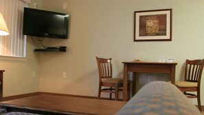 Hotellihuone, jossa on sänky, seinälle asennettu televisio, pieni pöytä tuoleineen ja seinällä oleva kehystetty kuva.