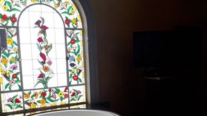 A bathroom with a white bathtub, a large stained glass window with floral designs, and a television on the wall. - Villas Princess Hotel (Mexico City)