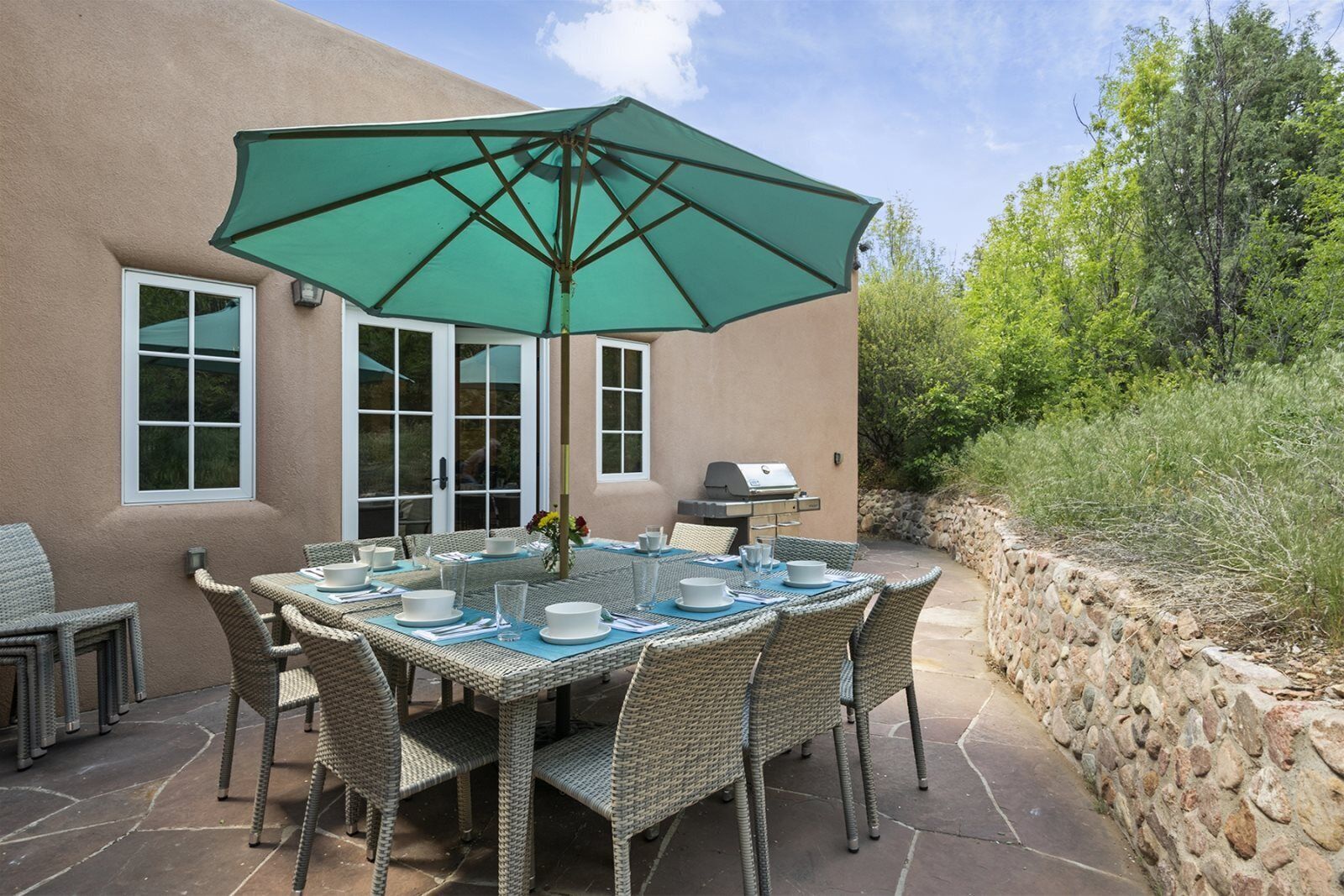 Eine Terrasse im Freien mit Esstisch, umgeben von Korbstühlen, einem grünen Sonnenschirm und einem Grill.