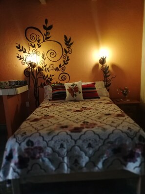 A bedroom with a patterned bedspread, a decorative wall mural, and two wall-mounted lamps. - Casa de Huespedes Independencia (San Miguel de Allende)