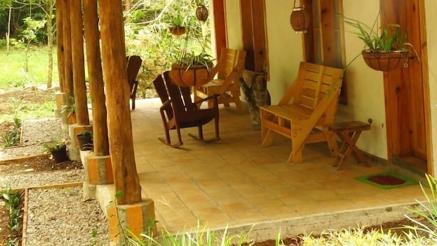 A covered outdoor seating area with wooden chairs and tables, surrounded by potted plants and a stone pathway. - Ecofinca Luna del Puente (Santa Cruz de Yojoa)