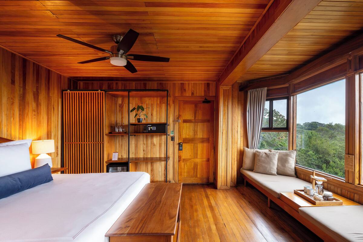 Une pièce en bois comprenant un lit, un banc, un ventilateur de plafond et de grandes fenêtres donnant sur l’extérieur.