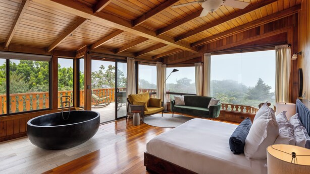 Une chambre spacieuse avec un grand lit, une baignoire noire et un balcon offrant une vue sur la forêt.