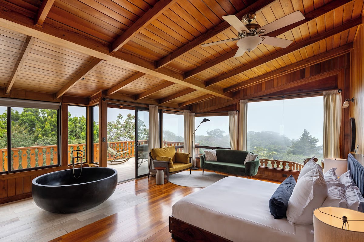 Une chambre spacieuse avec un grand lit, une baignoire noire et un balcon offrant une vue sur la forêt.